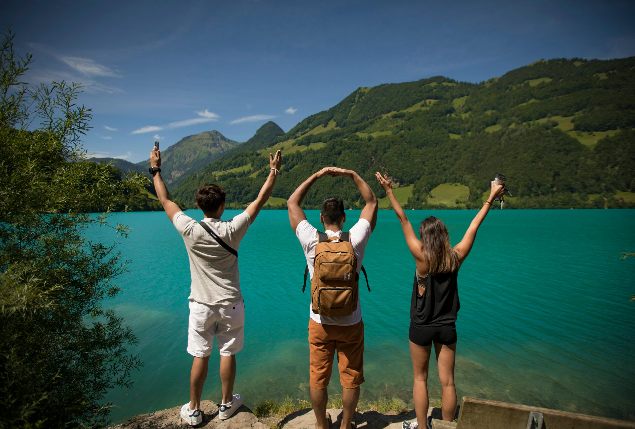 Three young adults celebrating by a vibrant turquoise lake surrounded by lush hills.